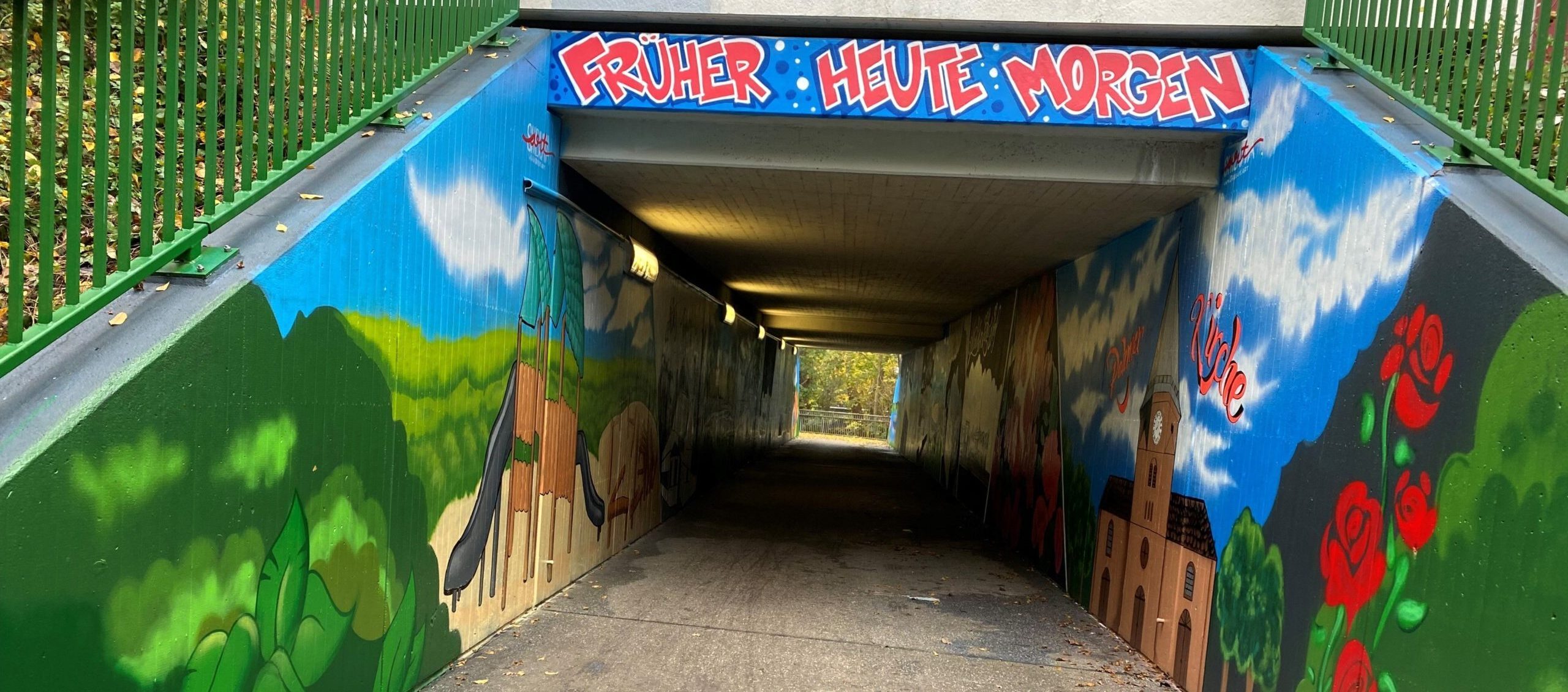 Graffiti-Kunst in einem Unterführungstunnel, der mit bunten Wandmalereien von Blumen, Bäumen und einer Kirche dekoriert ist. Der Tunnel ist gut beleuchtet und bietet einen einladenden Zugang zu einem Parkbereich.