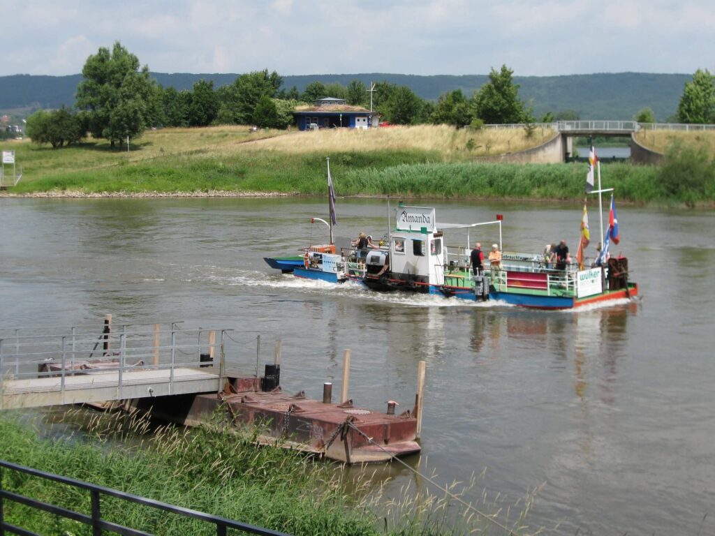 Fähre auf einem Fluss mit Passagieren, die zur anderen Uferseite transportiert werden. Im Hintergrund grüne Wiesen und Bäume, während das Wasser ruhig fließt. Eine kleine Anlegestelle ist im Vordergrund sichtbar.