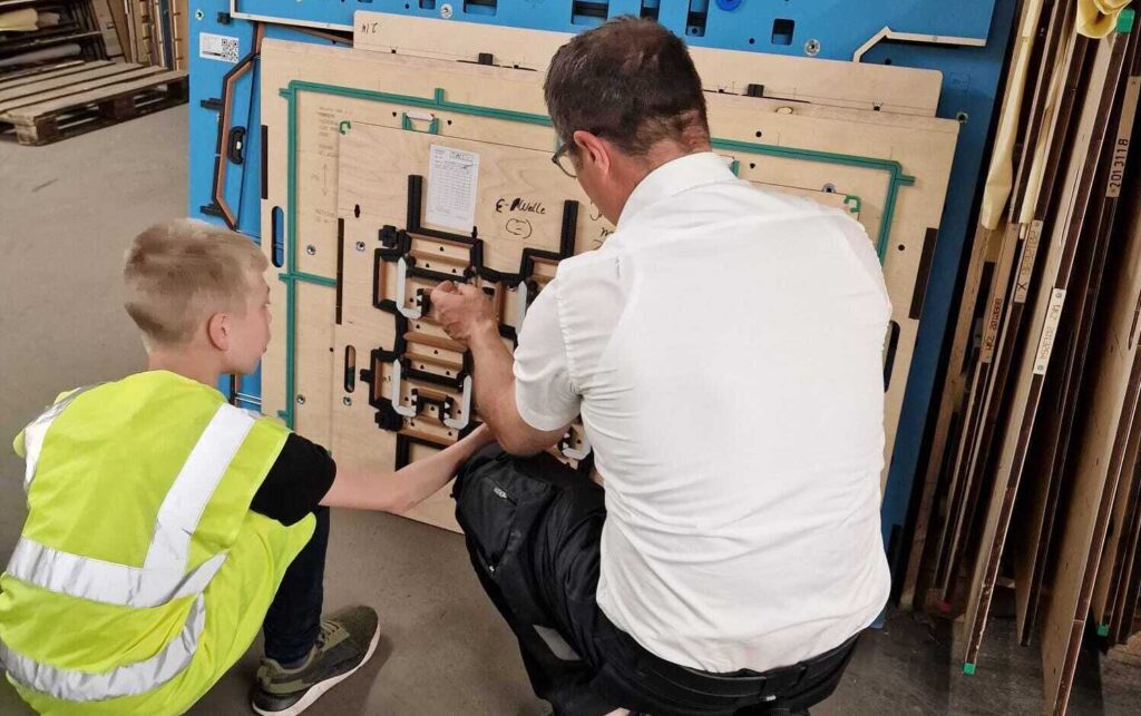 Ein Erwachsener und ein Kind arbeiten gemeinsam an einem technischen Gerät in einer Werkstatt. Der Erwachsene zeigt dem Kind, wie man verschiedene Komponenten montiert. Beide sind konzentriert und tragen Sicherheitskleidung, während sie an einem blauen Tisch mit Holzplatten und Werkzeugen arbeiten. Im Hintergrund sind weitere Materialien sichtbar, die auf eine aktive Werkstattumgebung hinweisen.